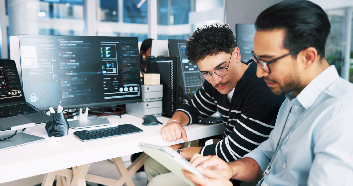 Two men looking at a tablet with a computer in the background